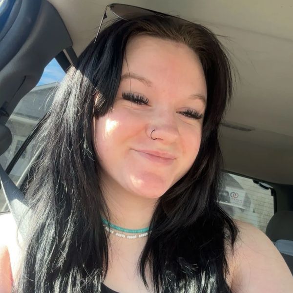 Smiling woman with long black hair inside a car wearing a seatbelt and sunglasses on her head.