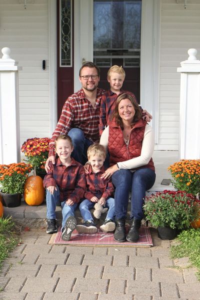 A family of five in coordinated plaid outfits sits on a porch surrounded by pumpkins and flowers.