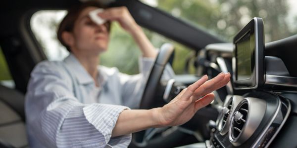 Woman wiping forehead and reaching for car dashboard controls, looking stressed.