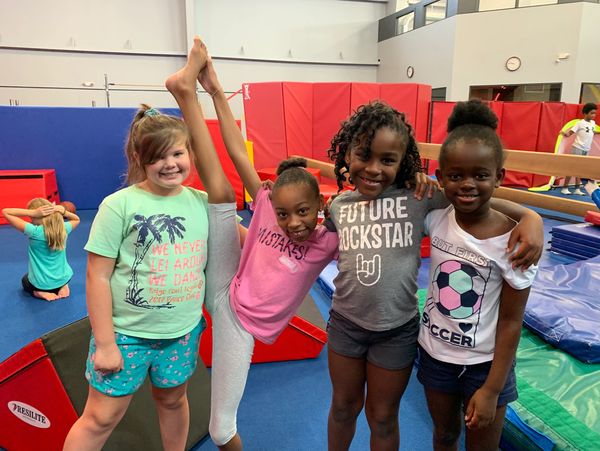 Four smiling girls posing in a gym with colorful mats and equipment.