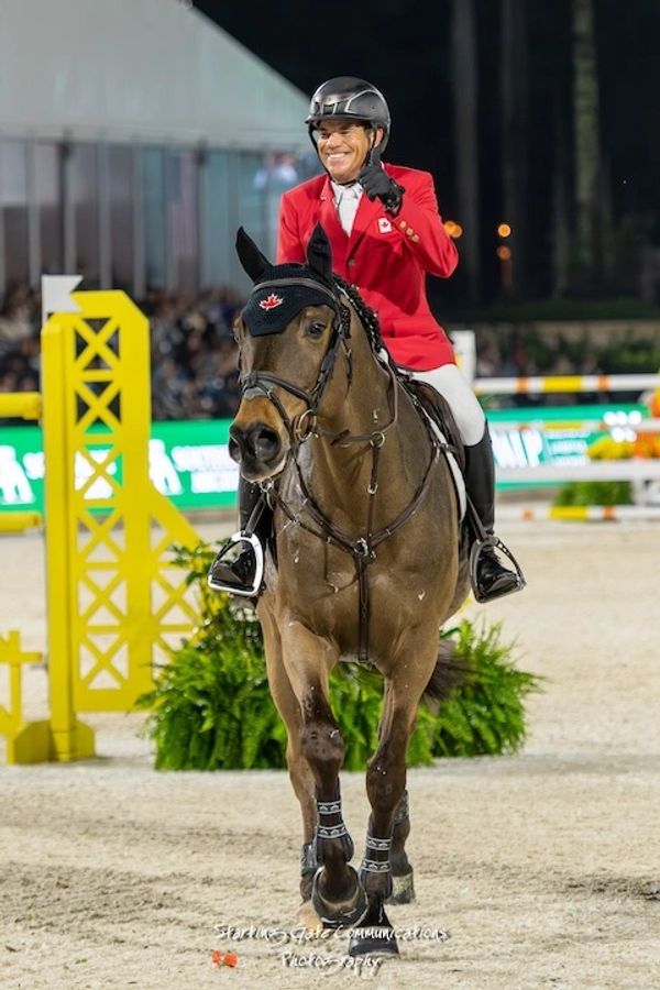 Smiling equestrian in red jacket on horse giving thumbs up during competition.