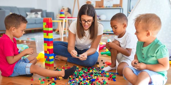 Handsome young teacher and little children playing with building blocks around a lot of toys at kind