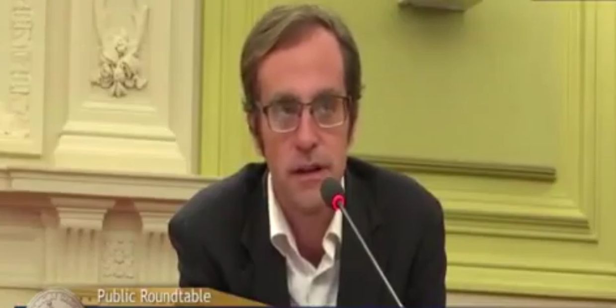 Man in glasses speaking at a public roundtable event in a formal setting.