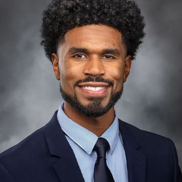 Representative Jesse Johnson wearing a navy blue suit and tie, smiling, behind a grey background