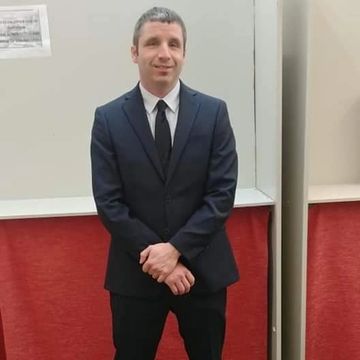 Mackenzie is standing, wearing a dark suit and tie, in front of a white and red wall