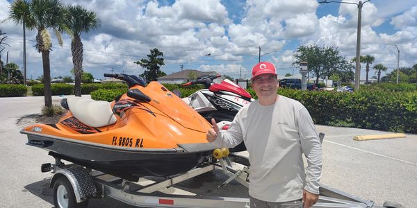 Man giving thumbs up next to two jet skis on a trailer in a sunny parking lot.
