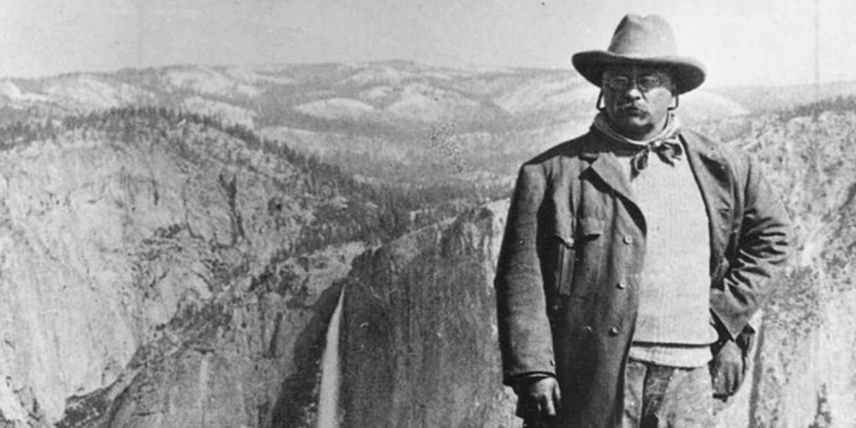 Historical black-and-white photo of a man in a hat near a waterfall in a mountainous area.