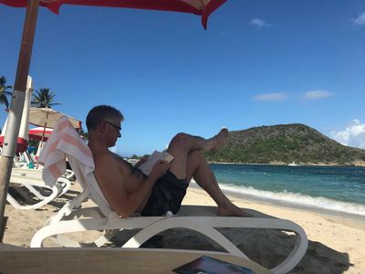 A man reading on a beach chair under an umbrella by the sea.