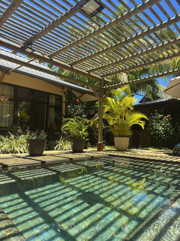 Sunlit backyard pool with pergola casting striped shadows and lush greenery.