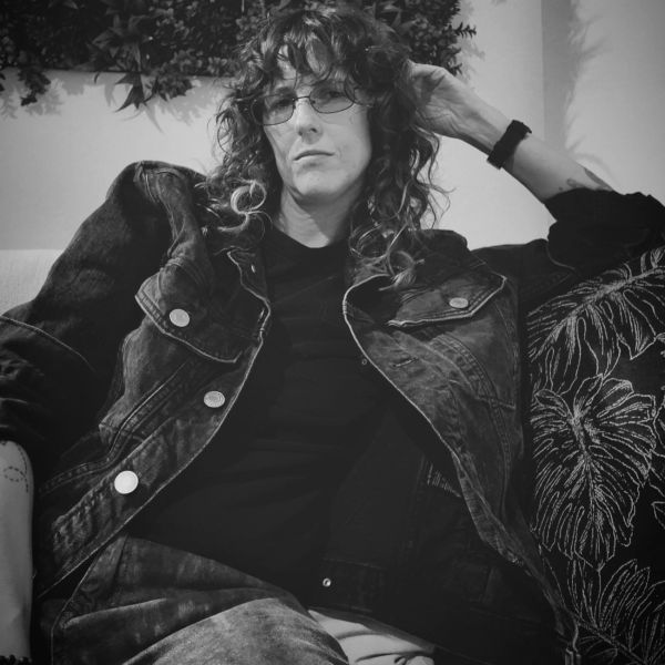 Person with curly hair and glasses wearing a denim jacket, lounging on a sofa.