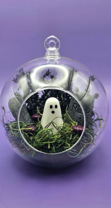 A ghost figurine inside a glass terrarium with moss and tombstones.