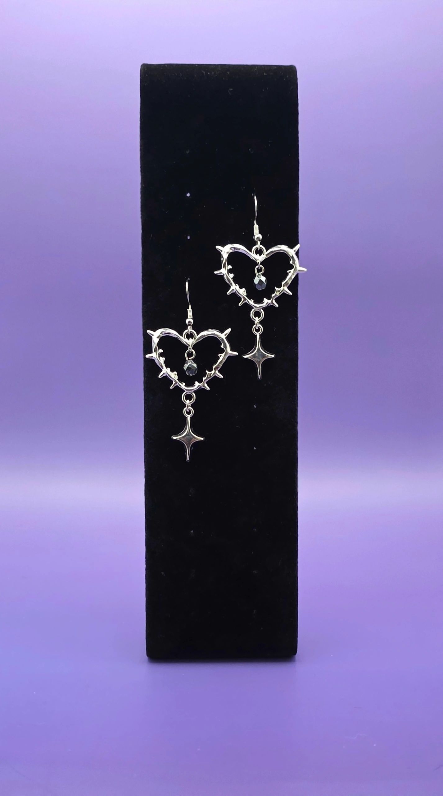 Silver heart-shaped earrings with dangling star charms on a black stand.