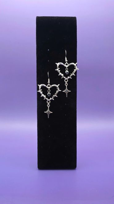 Silver heart-shaped earrings with dangling star charms on a black stand.