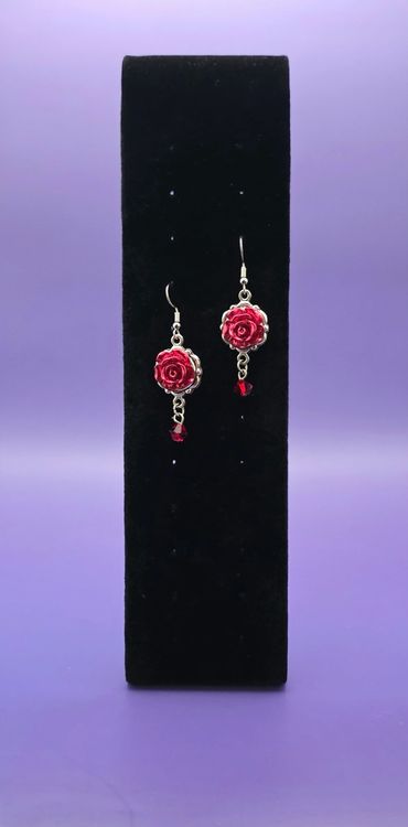Red rose earrings with dangling beads on a black display against a purple background.