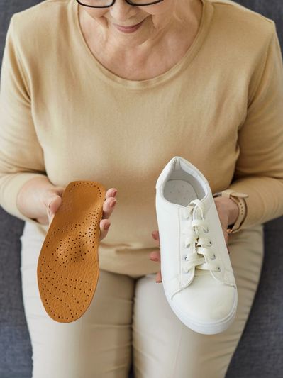 An elderly woman holding a white sneaker and a brown orthopedic insole.