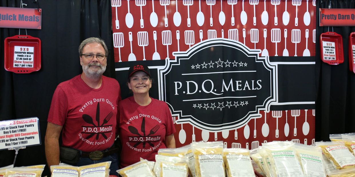 Two vendors wearing matching red shirts at P.D.Q. Meals booth with food packages.