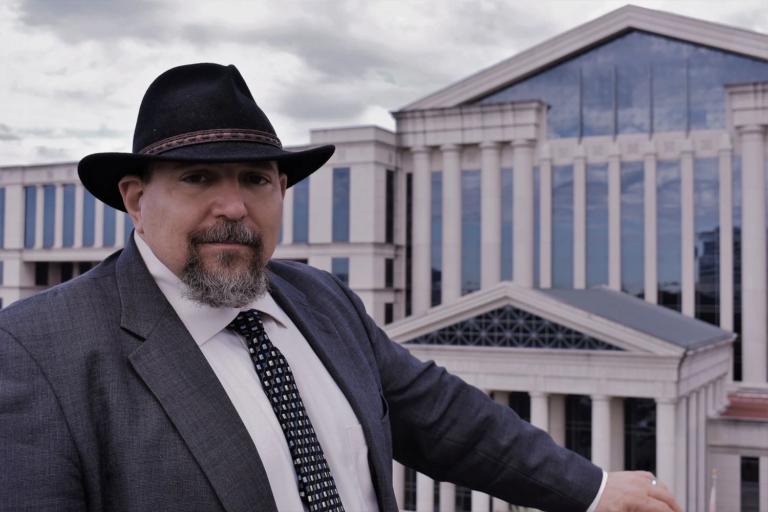 Man in a suit and hat poses confidently in front of a grand building.