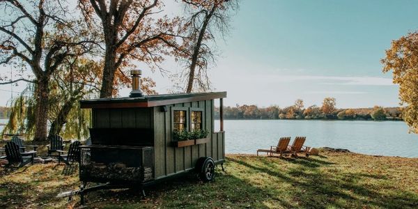 A cozy tiny cabin on wheels by a lakeside with chairs and autumn trees.