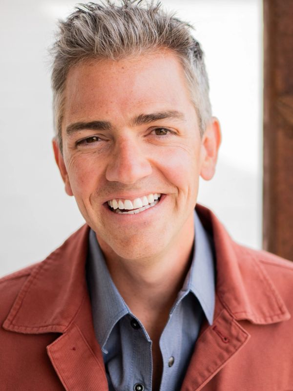 Smiling man with gray hair wearing a rust-colored jacket and blue shirt.