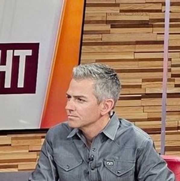 Man with gray hair in a gray button-up shirt on a TV set.