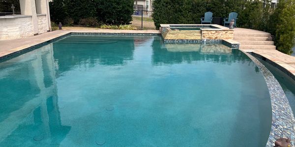 Clear outdoor swimming pool with attached hot tub and patio chairs.