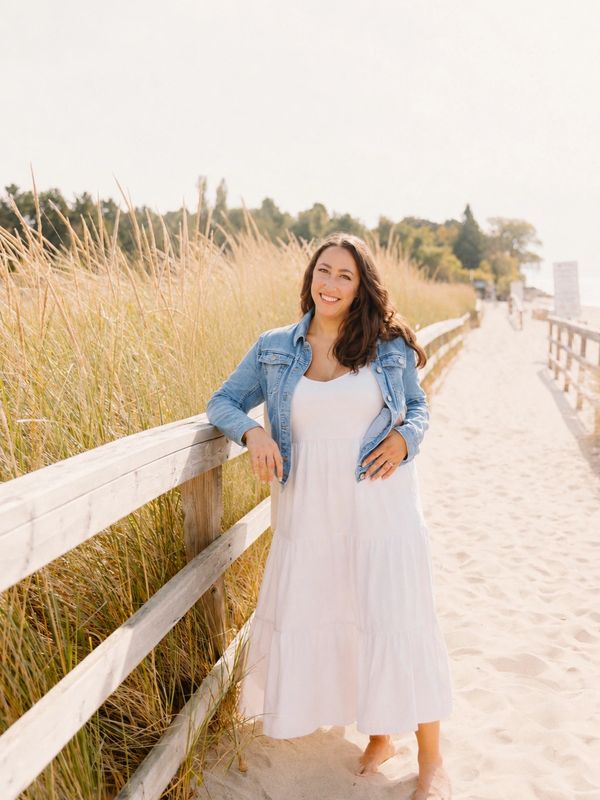 Woman in a white dress and denim jacket standing by a wooden fence on a sandy path.