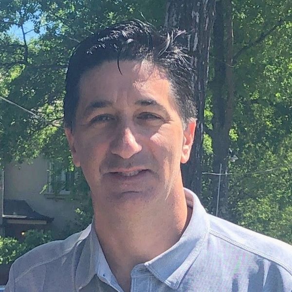 Smiling man outdoors with trees and sunlight behind him.