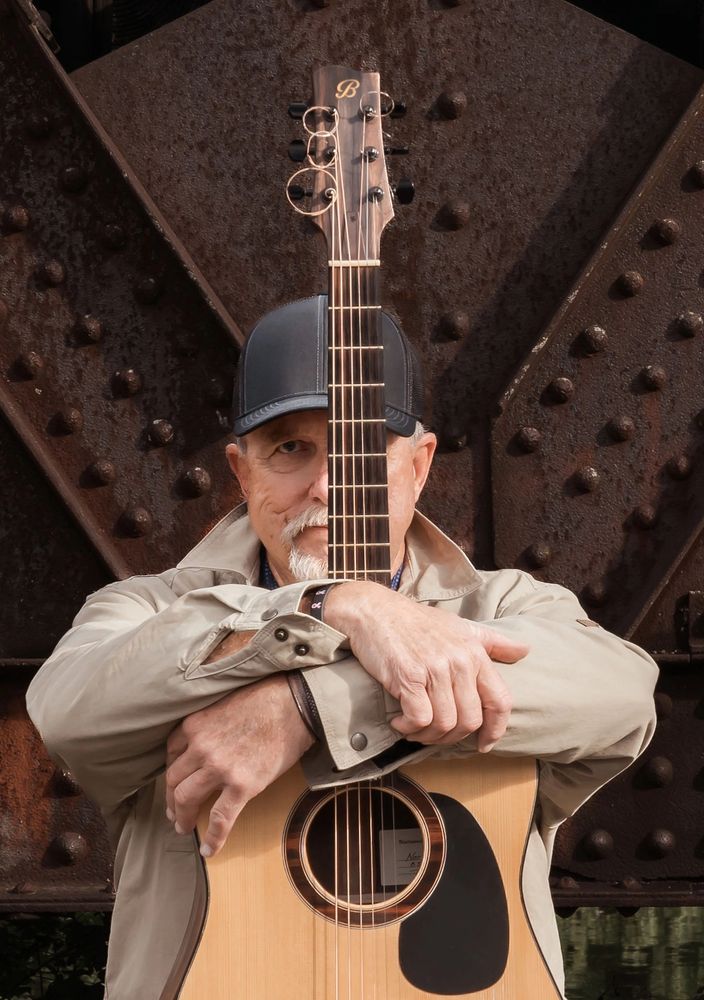 Man hugging an acoustic guitar in front of a rusty metal background.