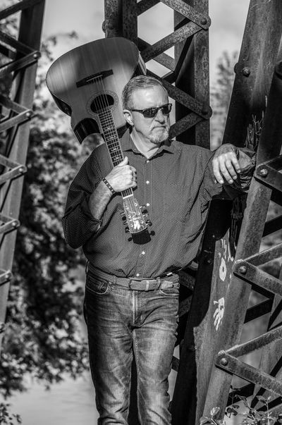 Man with guitar leaning on a metal structure in black and white.