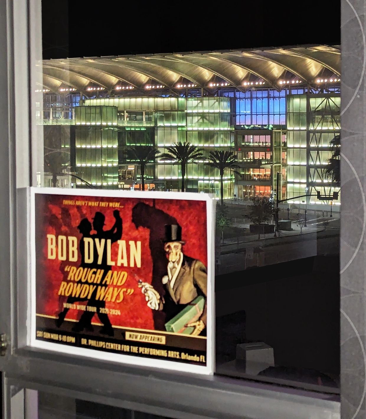 Bob Dylan concert poster displayed on a window with a modern illuminated building at night.