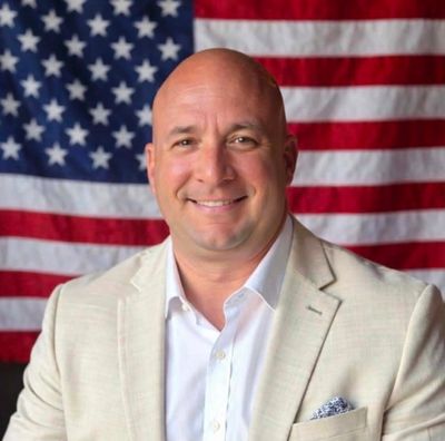 Smiling man in beige jacket with American flag background.