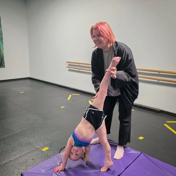 An instructor helps a young girl with a handstand stretch in a gym.
