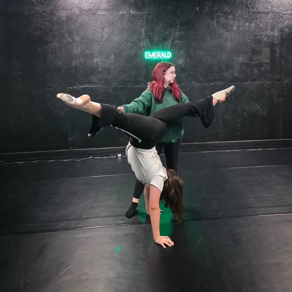 One dancer supports another doing a handstand split in a dance studio.