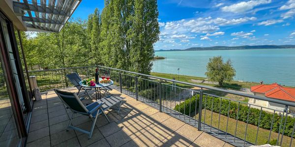 Balcony with chairs and a table overlooking a serene lake and trees under a blue sky.
