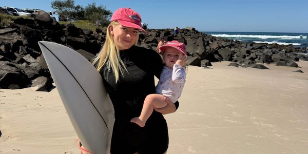 Woman holding child and surfboard on rocky beach under blue sky.