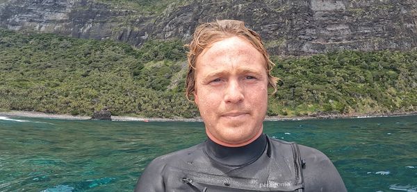 A man in a wetsuit stands near clear blue water with a rocky, green cliff in the background.