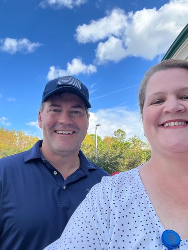 A smiling man and woman outdoors under a blue sky with clouds.
