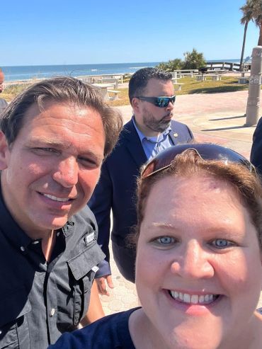 Two people smiling for a selfie near a beach with a man in sunglasses in the background.