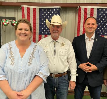 Three people posing in front of American flags, one wearing a cowboy hat and sheriff badge.