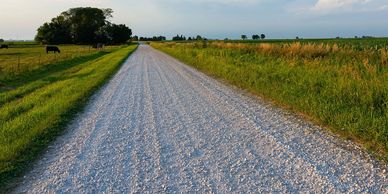 A gravel road stretches through green fields under a blue sky.
