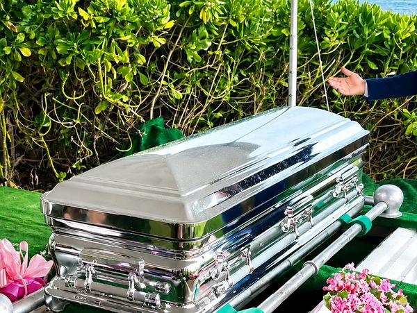 A shiny silver coffin at an outdoor funeral with flowers nearby.