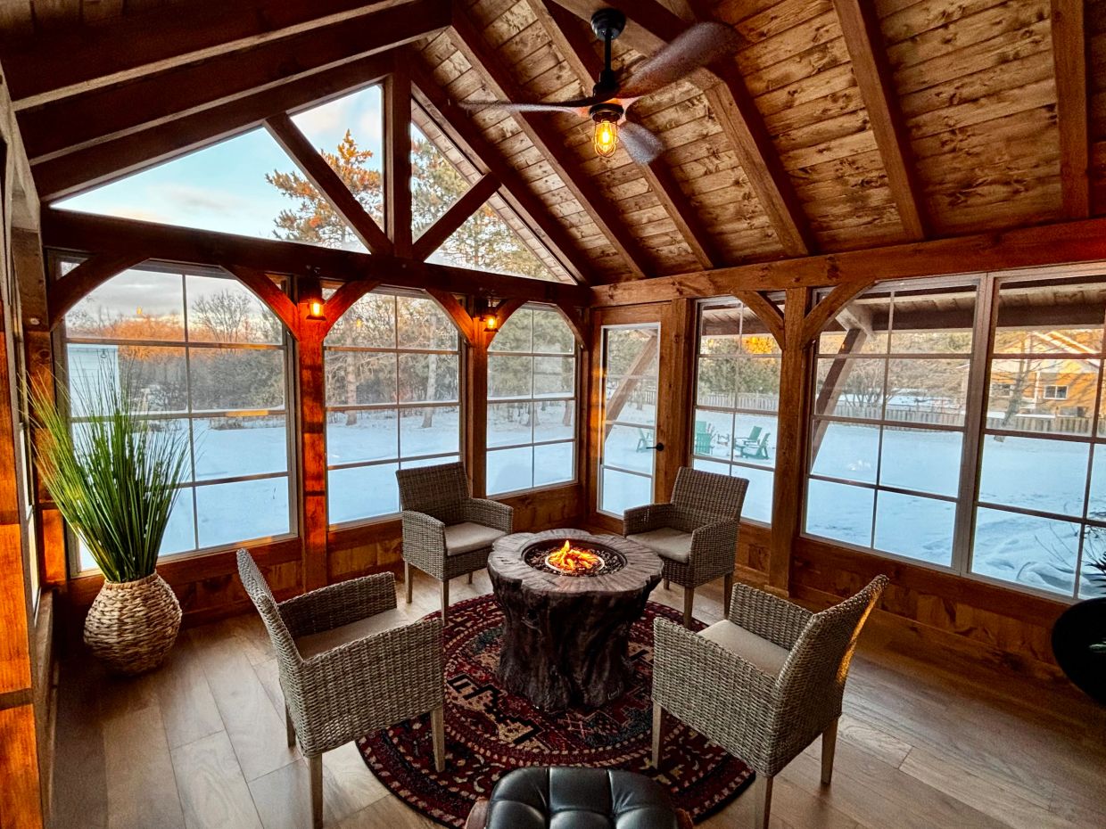 Cozy wooden sunroom with wicker chairs around a fire table and snowy outdoors.
