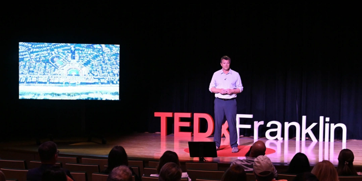 A speaker presents at TEDxFranklin with a city aerial view on screen.