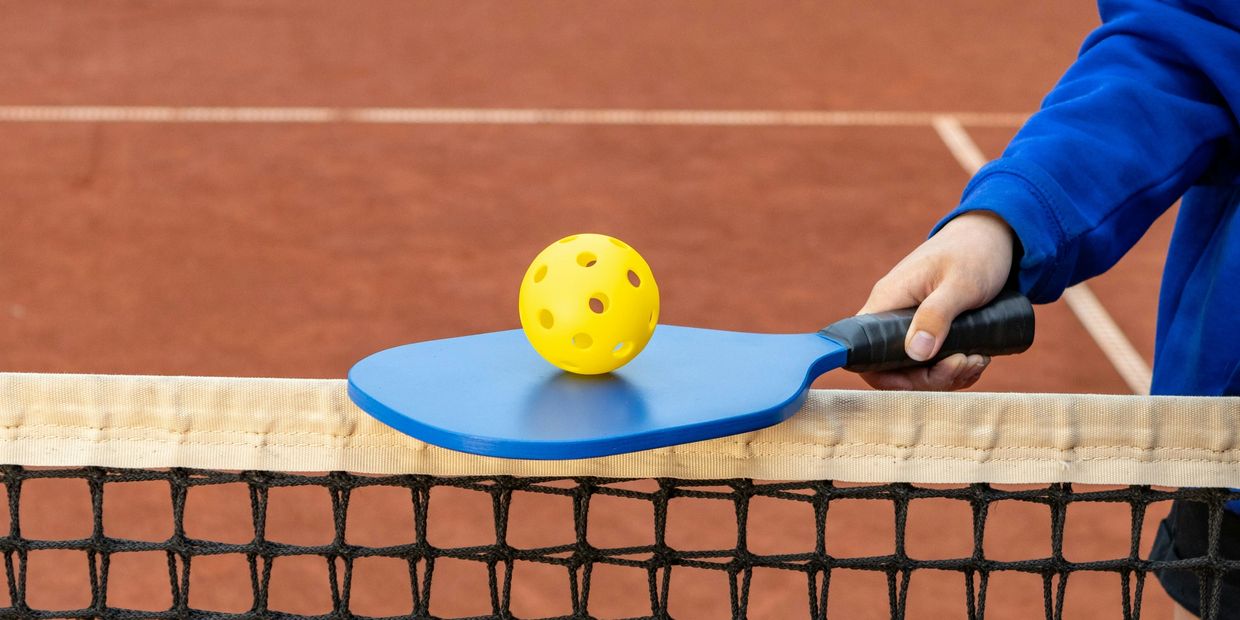 A hand holding a paddle with a yellow ball over a net.