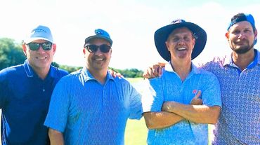 Four men smiling outdoors on a sunny day, dressed casually in golf attire.
