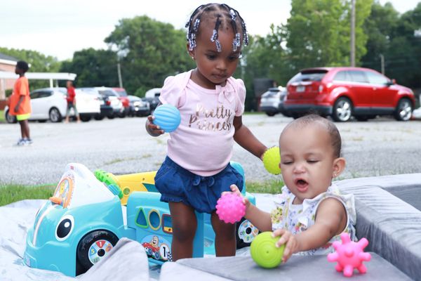 Two toddlers playing with colorful textured balls near a toy car outdoors.