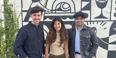 Three friends smiling, standing against a black and white mural wall with greenery on the side.