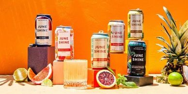 Colorful June Shine hard kombucha cans arranged with fresh fruits against an orange background.
