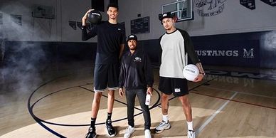 Three men posing on a basketball court, two holding basketballs.