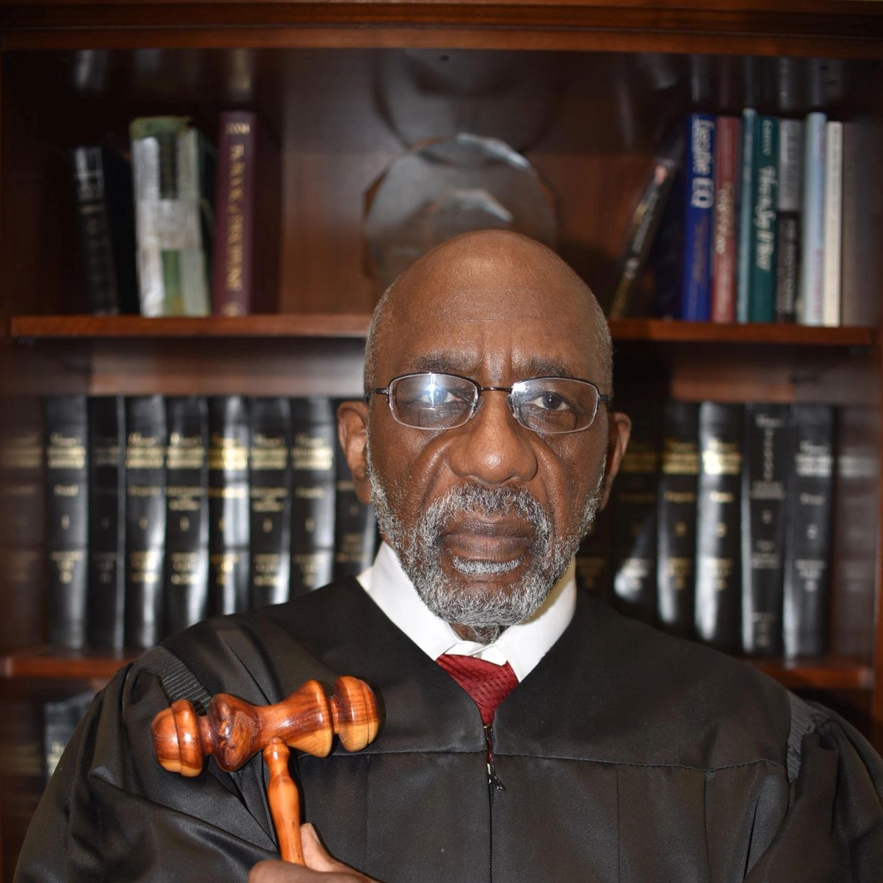 Judge in black robe holding a gavel, standing in front of a bookshelf filled with legal books.